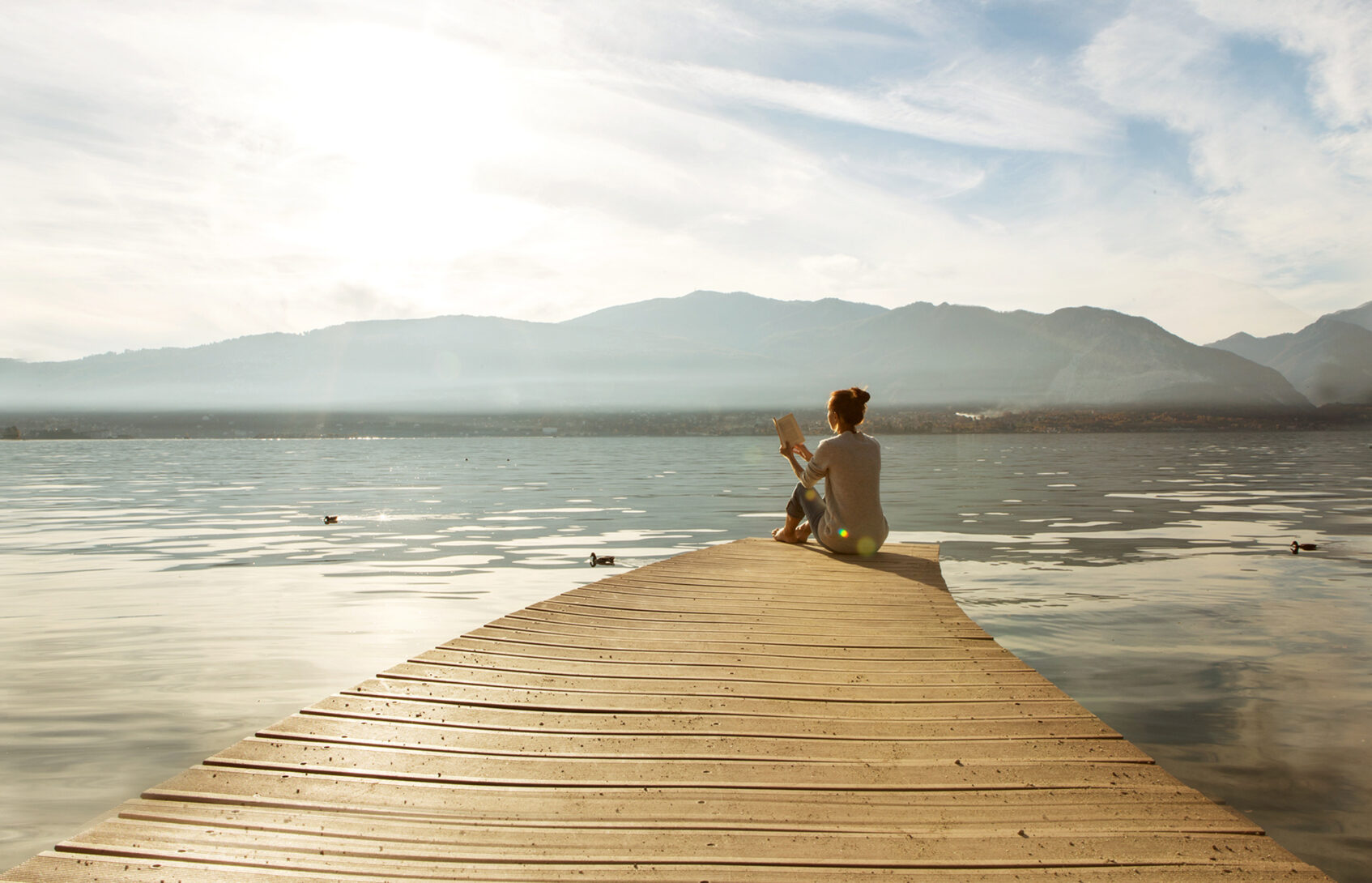 A woman sits reading a book on a dock on a lake