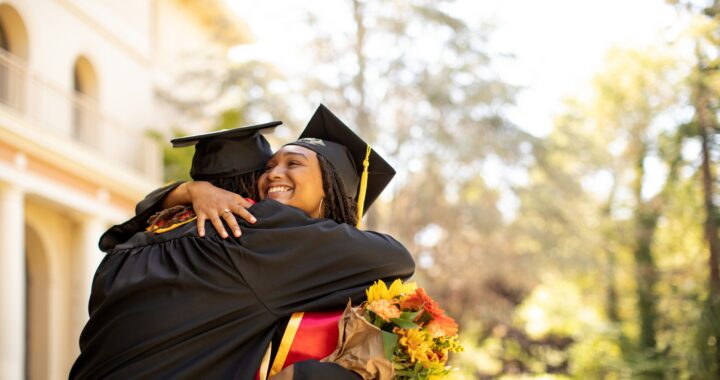 Two students hug while wearing graduation caps and gowns.