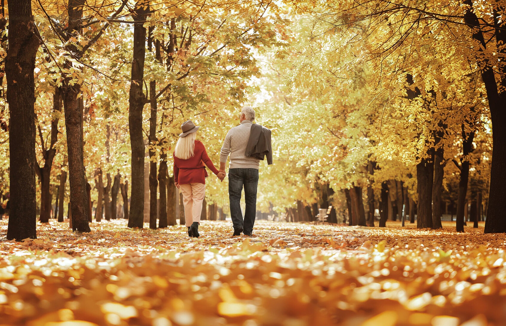 An older couple walks along a trail covered in leaves and surrounded by colorful trees.